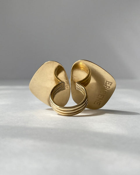 Product shot of the back of a large brass ring consisting of two side by side abstract geometric shapes and the EPICENE logo stamped into the metal, with wide brass band shown on a white background
