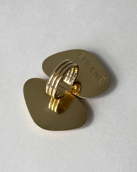Product shot of the back of a large brass ring consisting of two side by side abstract geometric shapes and the EPICENE logo stamped into the metal, pictured face down on a white background