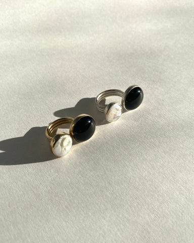 One brass ring and one silver ring, both open, each with a round bezel-set black onyx and cream freshwater pearl photographed in the sun on a white background