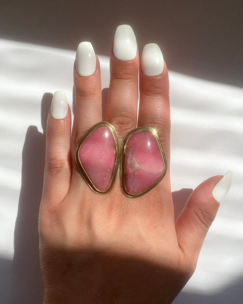 Large brass statement ring with two abstract pieces of pink rhodonite stone shown on hand with long white nails photographed in the sun