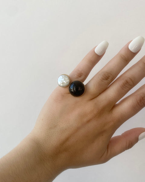 Brass pinky ring with a round bezel-set black onyx and round cream freshwater pearl photographed on a hand with long white nails on a white background