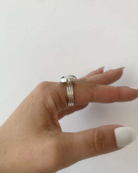 Side view of silver wide band ring with  bezel-set freshwater pearl sticking out on top  photographed on hand with long white nails and a white background
