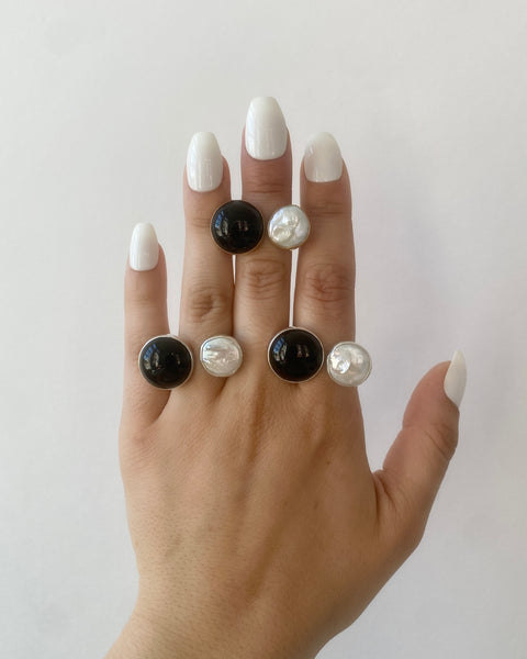 Three rings, each with a round bezel-set black onyx and cream freshwater pearl photographed on a hand with long white nails on a white background
