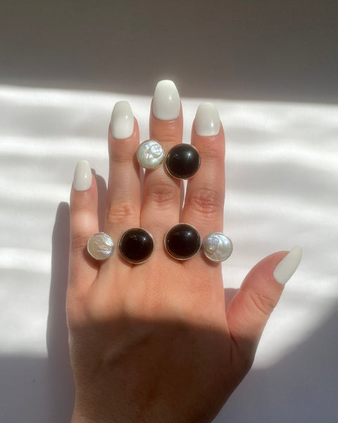 Three rings, each with a round bezel-set black onyx and cream freshwater pearl photographed on a hand with long white nails in the sun on a white background 
