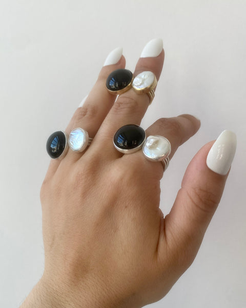 Three-quarter view of three rings, each with a round bezel-set black onyx and cream freshwater pearl photographed on a hand with long white nails on a white background