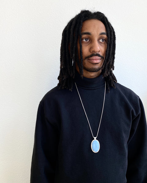 Brandon, a handsome stylish young man wears the long blue opal pendant  over his black sweatshirt as a long statement necklace